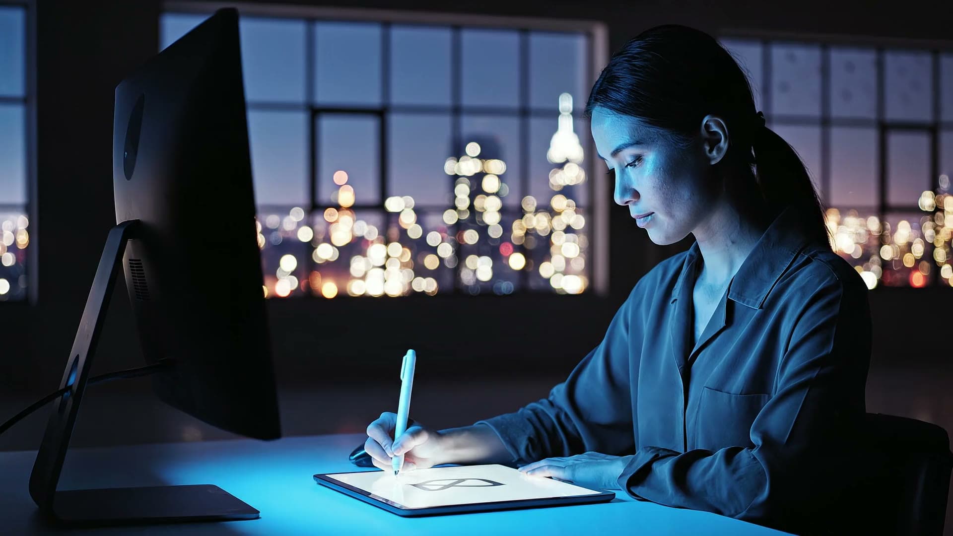 AI-generated still from the studio film: a designer at a desk at night sketching on a lit tablet next to a monitor, city lights blurred behind her