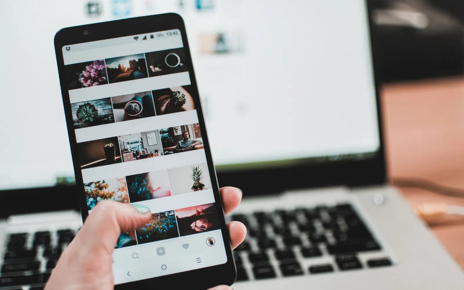 Une main tient un smartphone affichant un fil de réseau social devant un ordinateur portable