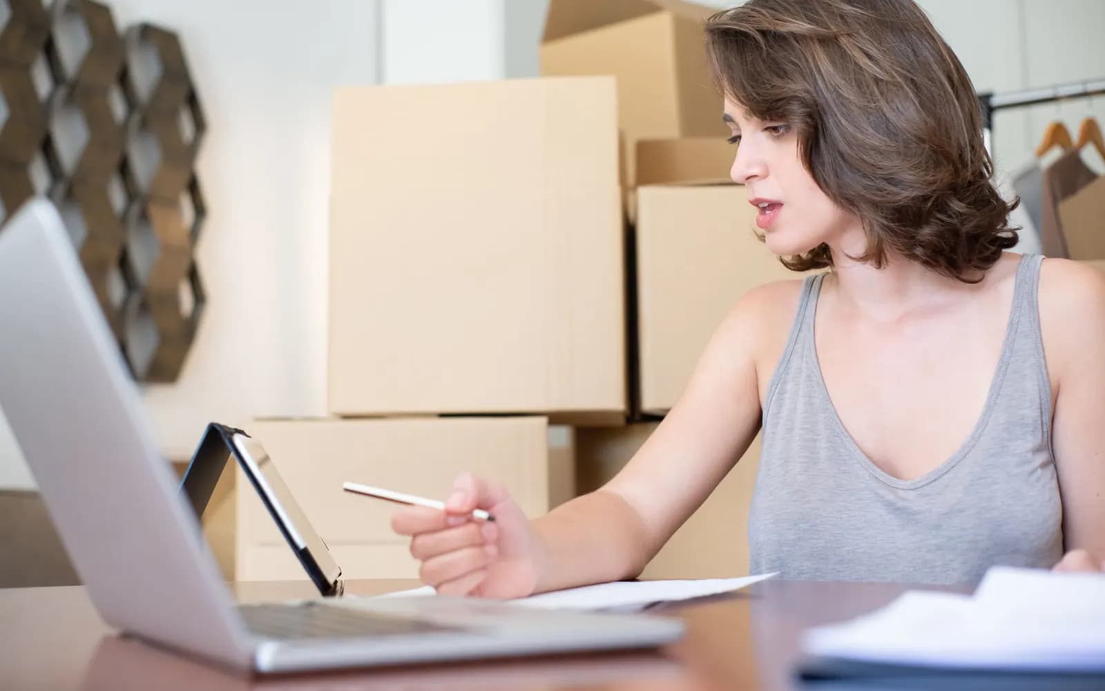 Une vendeuse prend des notes devant un ordinateur portable et une tablette, entre les cartons d’une boutique en ligne