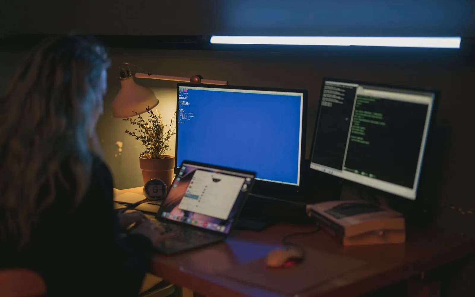 A developer works in a dim room in front of several screens with code and a terminal