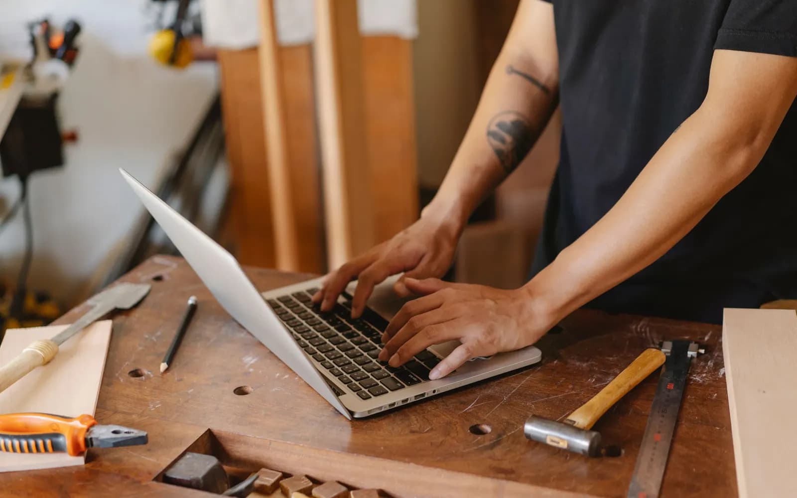 Un artisan tape sur un ordinateur portable à son établi, entouré d’outils de menuiserie